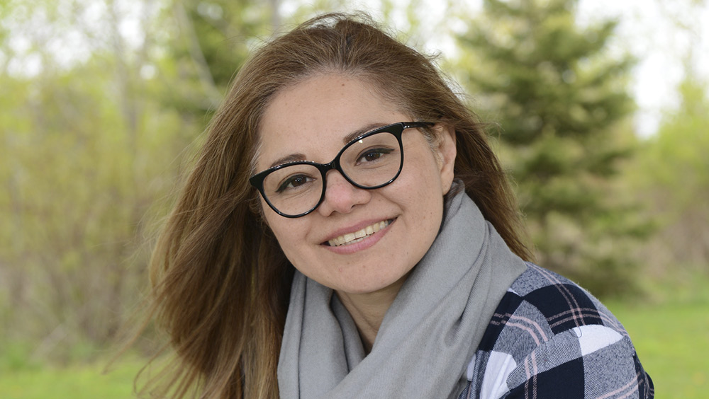 Photo of Vilma wearing black glasses, a plaid shirt and grey scarf posed outside in front of greenery. 