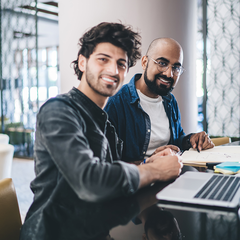 Two Middle Eastern man sitting side by side smiling to the camera, laptop and note book on the table.