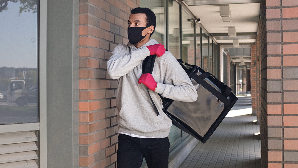 A young man standing near glass window carrying a delivery box on his back.